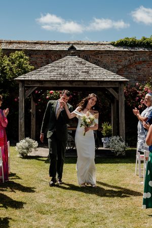 Newlywed couple walking down outdoor aisle after ceremony, bride holding bouquet, guests applauding.