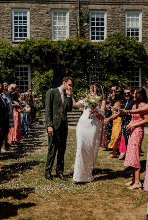 Newlywed couple walks through confetti shower outside stone manor house surrounded by cheering wedding guests.