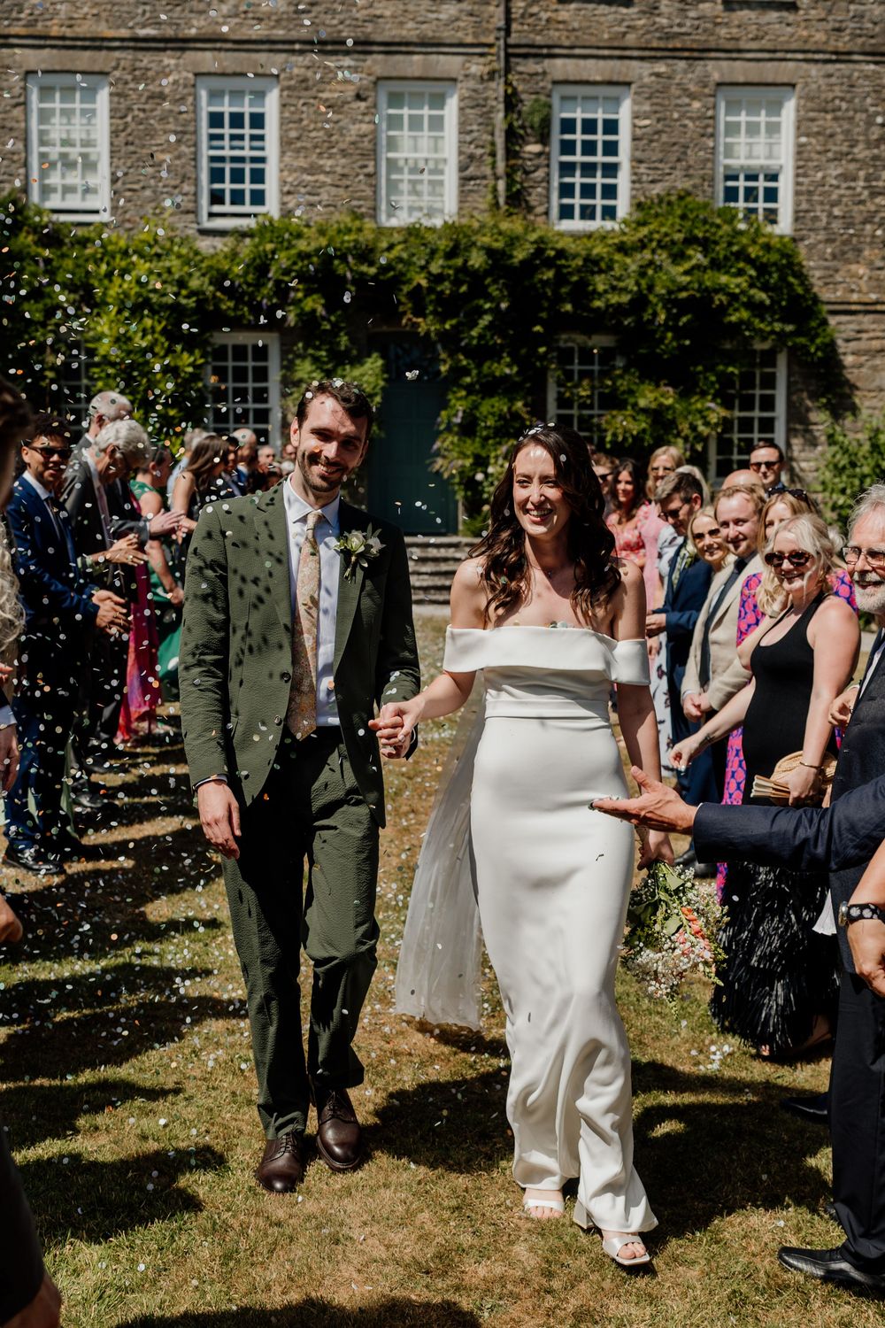 Bride and groom walk through confetti shower outside stone manor house on sunny wedding day.