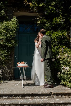 Bride and groom sharing a kiss outdoors by a teal door surrounded by lush greenery with a wedding cake on a white table.