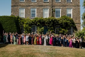 Large wedding group photo with guests cheering in front of an ivy-covered stone manor house on a sunny day.