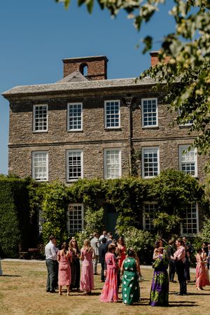 Wedding guests in colorful attire mingling outside an elegant ivy-covered stone country house under a clear blue sky.