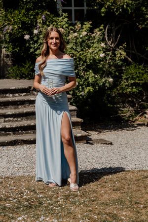 Woman in light blue off-shoulder slit gown holding champagne outdoors at a sunny garden wedding venue.