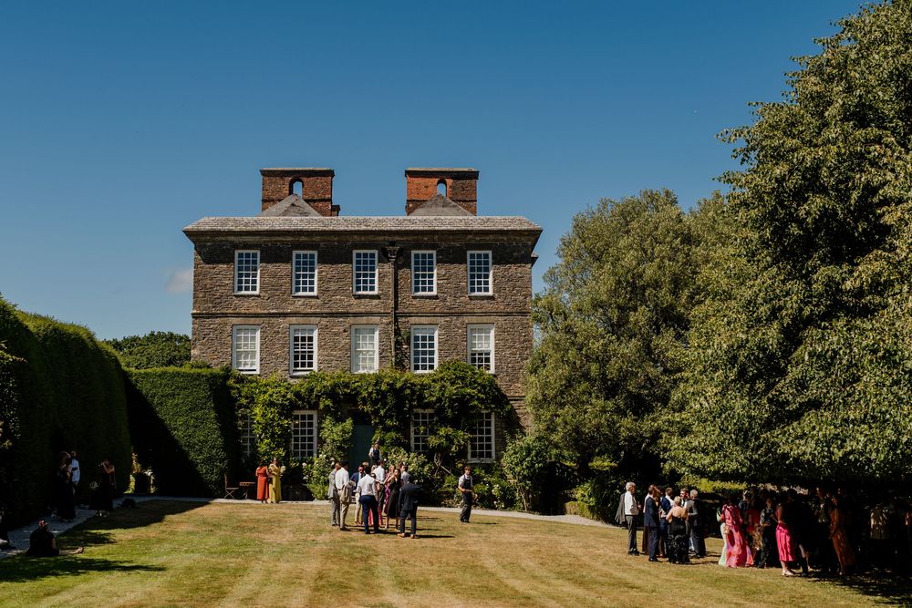 Elegant stone manor house wedding venue surrounded by manicured hedges and lawns with guests mingling outside.