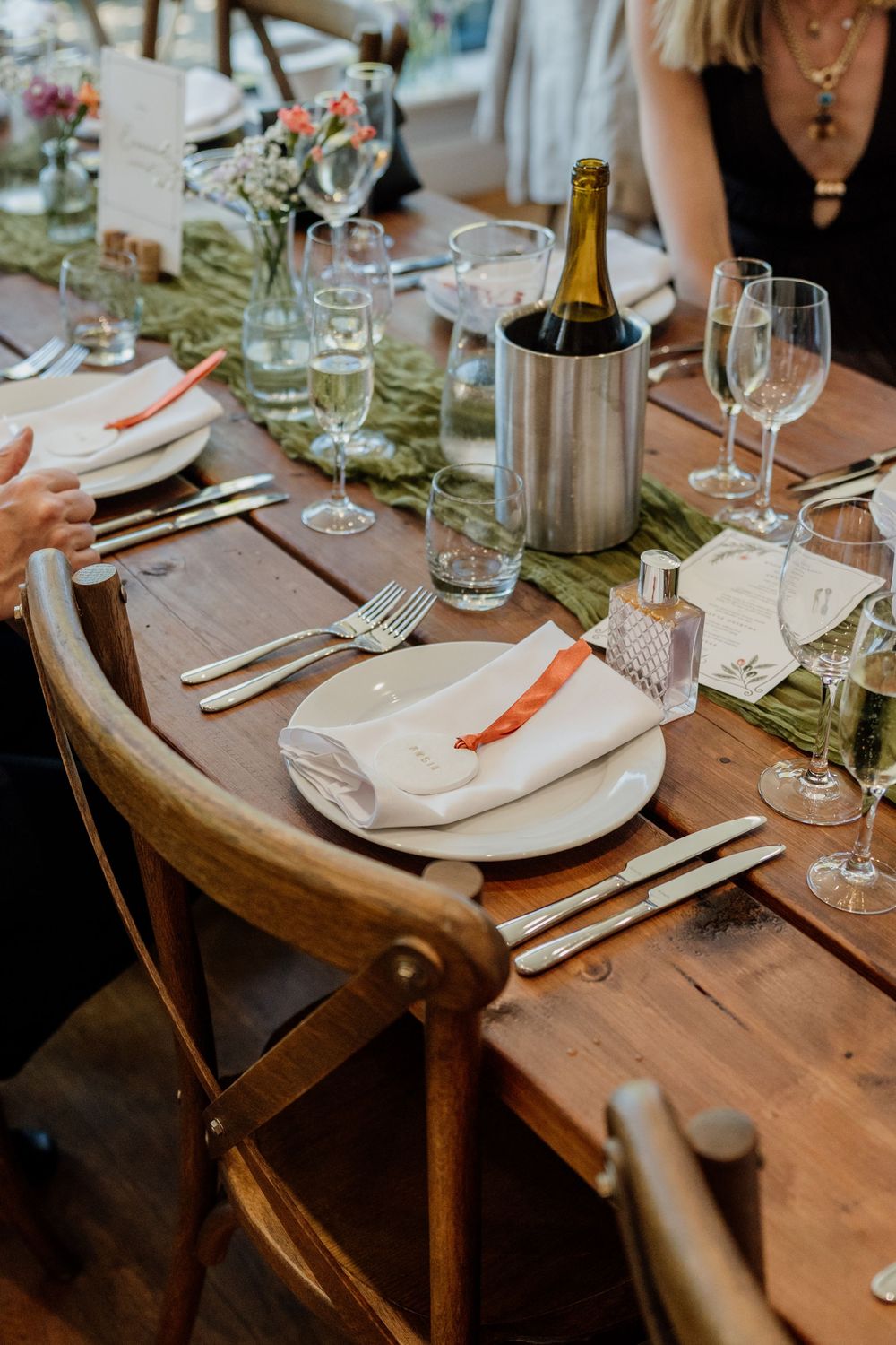 Elegant rustic wedding reception table with champagne bottle, crystal glasses, white plates, and orange napkins.