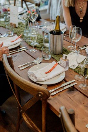 Elegant rustic wedding reception table with champagne bottle, crystal glasses, white plates, and orange napkins.