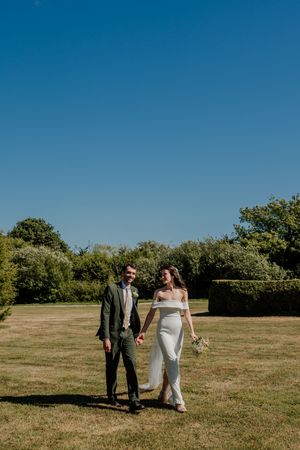 Bride and groom walk hand in hand across a sunny green lawn on their wedding day under a clear blue sky.