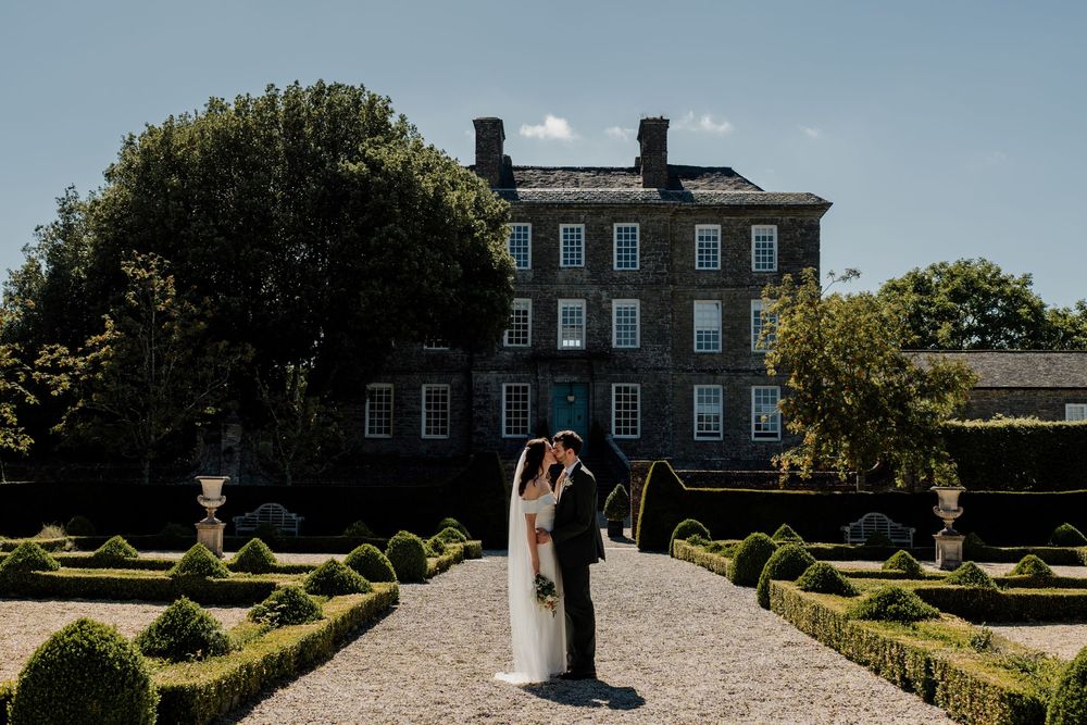 Bride and groom share a romantic kiss in a formal garden outside an elegant Georgian country house.