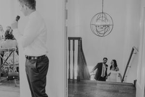Black and white wedding photo of bride and groom reflected in mirror while man stands in foreground.