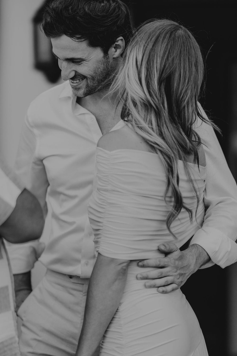 Black and white photo of smiling groom embracing bride in fitted white dress at wedding reception.