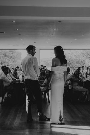 Bride and groom stand before wedding guests in elegant reception hall in black and white photo.