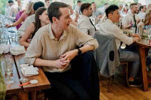 Man in beige linen shirt smiling at wedding reception, seated at long wooden table with guests.