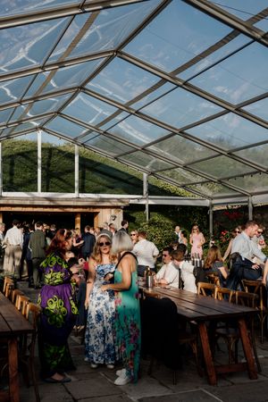 Wedding guests mingling under a clear marquee tent with wooden tables in a garden setting.