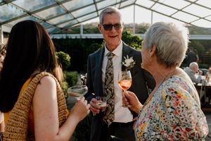 Smiling wedding guest in floral suit with boutonniere socializing with guests holding wine glasses outdoors.