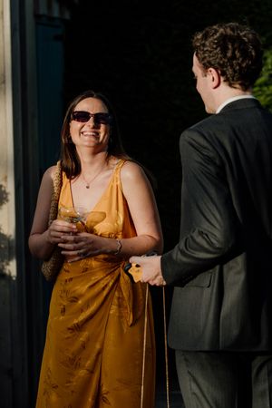 Laughing woman in golden yellow dress with sunglasses chatting with suited guest at wedding reception.
