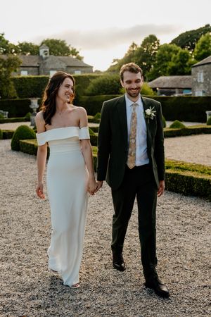 Bride in off-shoulder white gown and groom in dark suit holding hands in garden at golden hour.