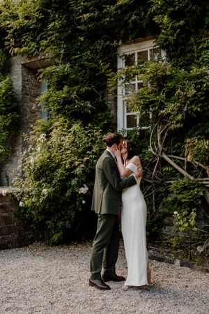 Bride and groom share romantic kiss beside ivy-covered stone wall on their wedding day.