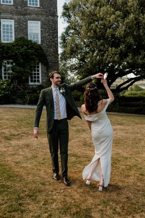 Bride and groom dancing outdoors on lawn beside stone manor house, groom in green suit twirling bride in white gown.