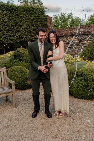 Joyful newlyweds popping champagne bottle in garden with string lights and manicured hedges at dusk.