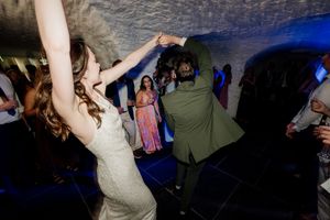 Bride and groom dancing joyfully at wedding reception, guests watching in background with blue ambient lighting.