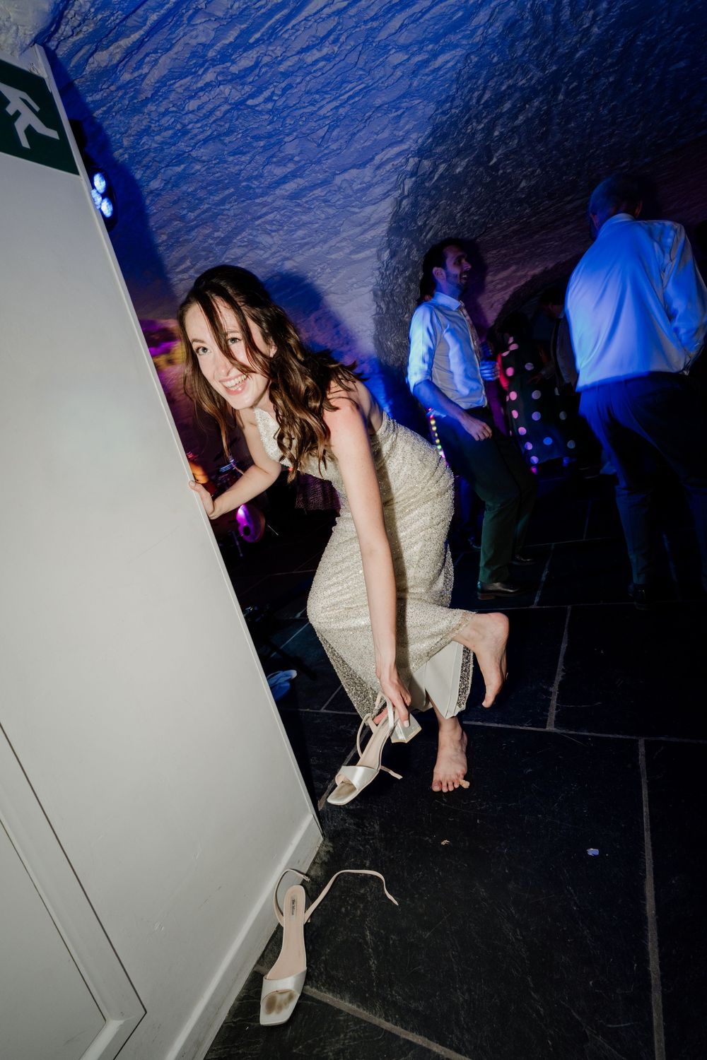 Bride laughing while removing her heels on a dark dance floor with colorful lights and guests dancing in the background.