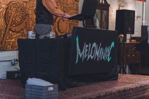 DJ Melomaniac performing behind a branded black table setup at an indoor event venue with decorative wall art.