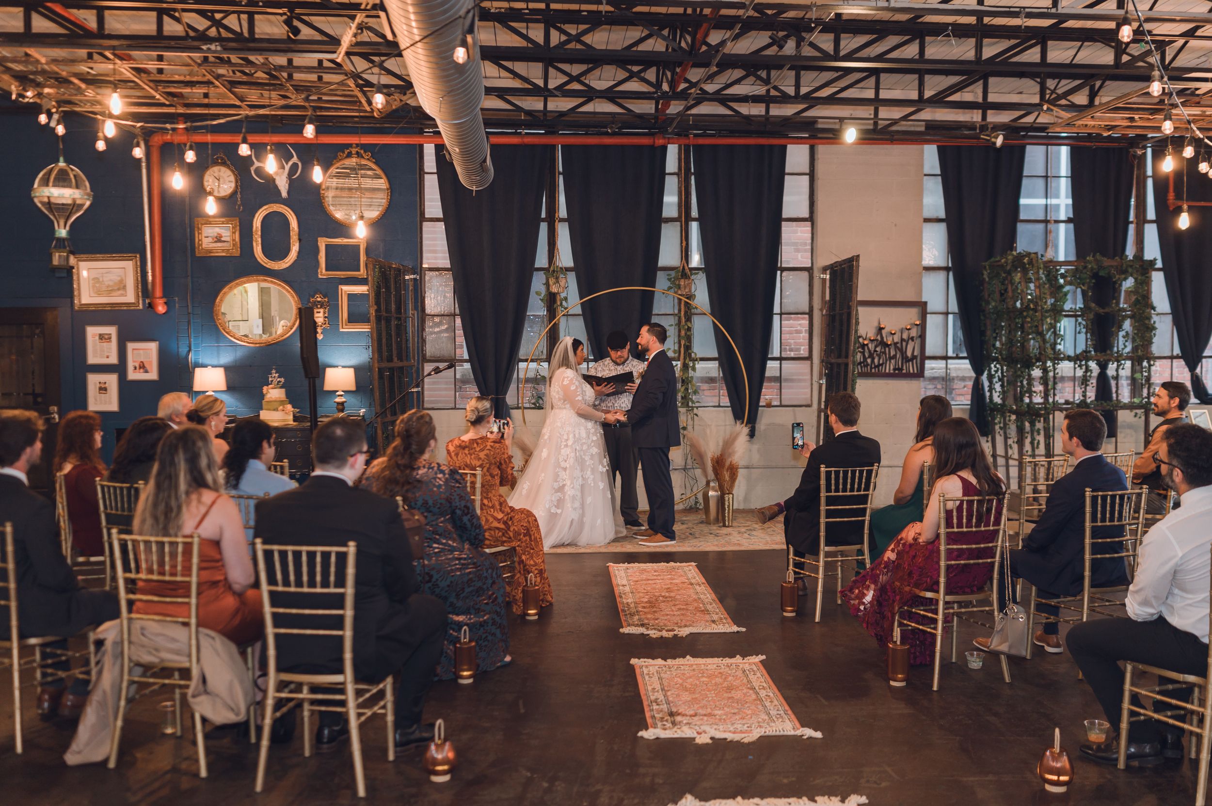 Indoor wedding ceremony with bride and groom at altar, guests seated in chiavari chairs, string lights and navy drapes.