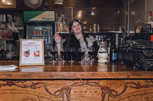 Smiling bartender waves behind a wooden bar with cocktail menu and wine glasses at a cozy restaurant.