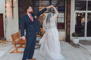 Groom in navy suit twirling bride in white lace wedding gown outdoors at rustic venue with string lights.