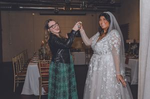 Bride in lace gown and veil dances joyfully with guest in leather jacket at wedding reception.