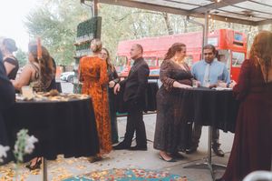 Elegantly dressed guests mingle at an outdoor evening event under a canopy with a red bus in the background.