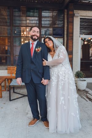 Smiling groom in navy suit and bride in floral lace gown holding hands at their wedding venue.