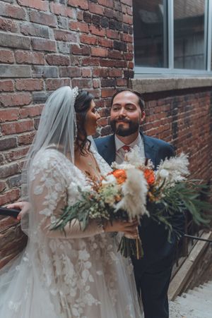 Bride and groom sharing a romantic moment against a rustic brick wall with a boho floral bouquet.