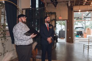 Groom in navy suit smiling during wedding ceremony while officiant reads from book in rustic industrial venue.