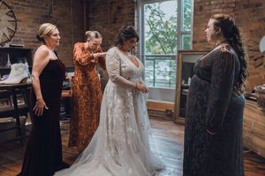 Bride in lace wedding gown being helped by three bridesmaids in elegant dresses inside a rustic brick venue.