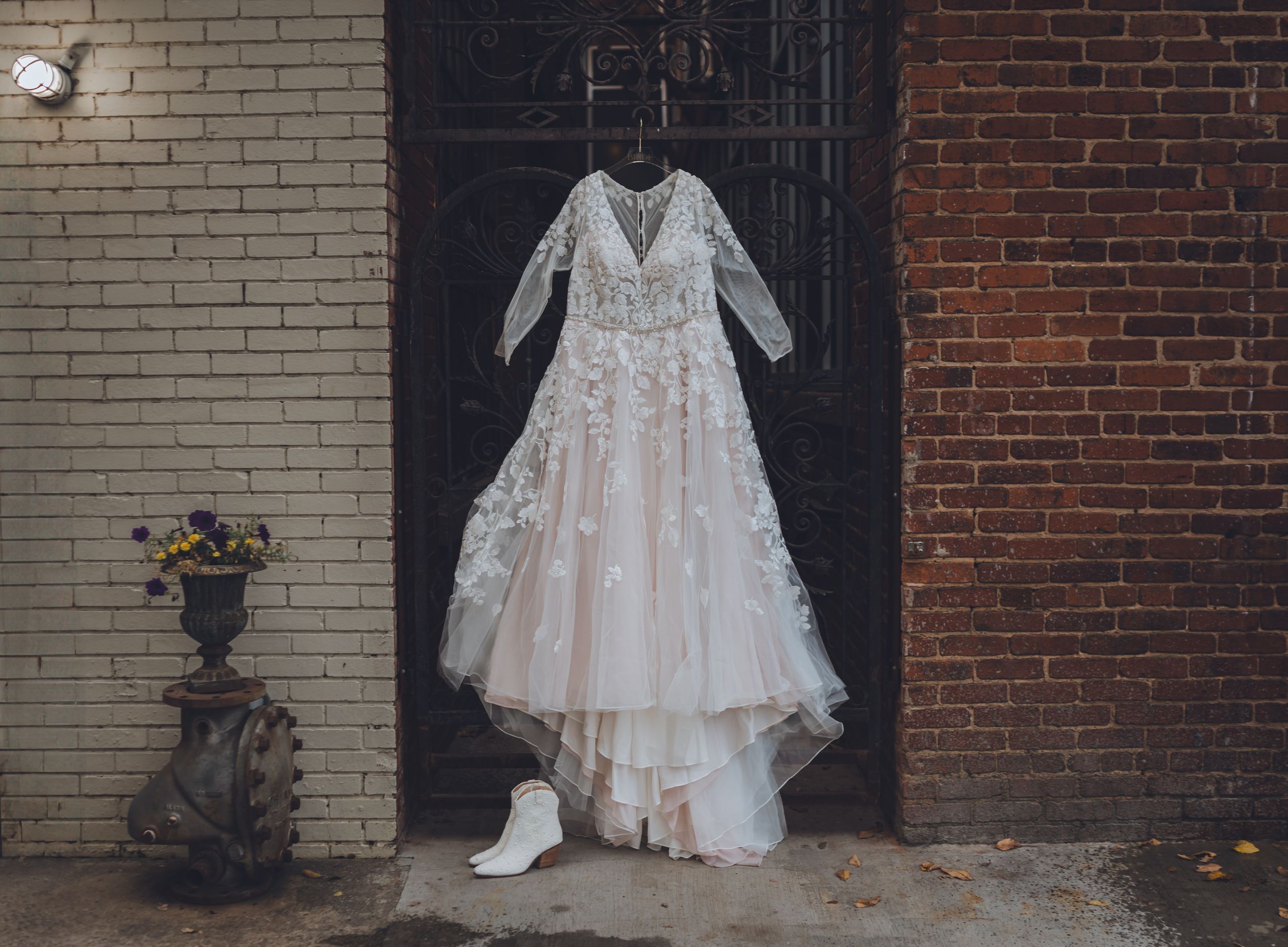 Elegant lace wedding dress with long sleeves and floral appliques hanging against a rustic brick wall with white bridal boots below.
