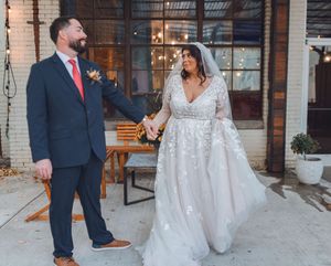 Bride in floral lace gown and groom in navy suit hold hands outside rustic brick venue on their wedding day.