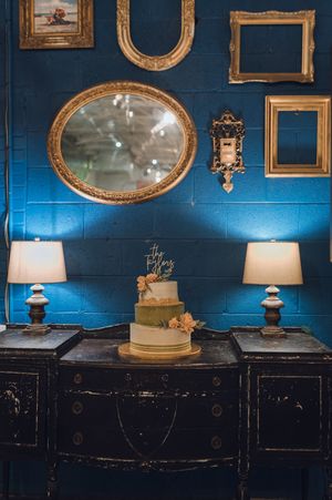 Three-tier wedding cake displayed on vintage black dresser against blue brick wall with gold mirrors and white table lamps.