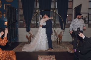 Bride and groom share first kiss under golden circular arch at indoor wedding ceremony with guests watching.