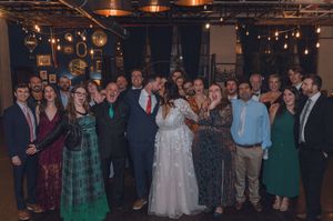 Wedding couple kissing surrounded by smiling guests in elegant attire at a warmly lit indoor reception venue.