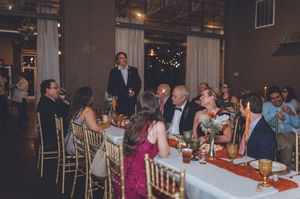 Guests laugh and celebrate at an elegant wedding reception dinner with gold chairs and string lights.