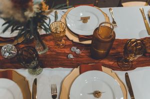 Elegant wedding table setting with white plates, gold chargers, amber glassware, and a wooden runner centerpiece.