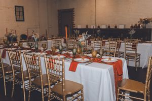 Elegant wedding reception hall with gold chiavari chairs, white tablecloths, orange runners, and floral centerpieces on long dining tables.