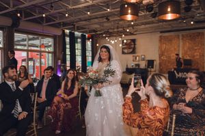 Bride walks down the aisle in a white gown holding a bouquet at an industrial-style wedding venue.