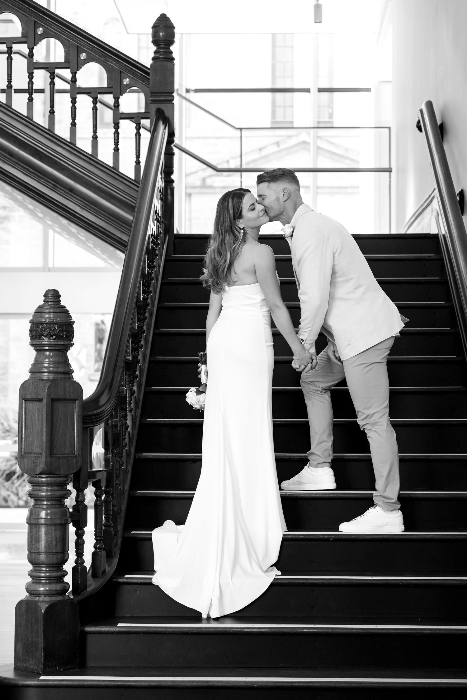 Black and white photo of a bride and groom sharing a kiss on elegant stairs, bride holding bouquet.