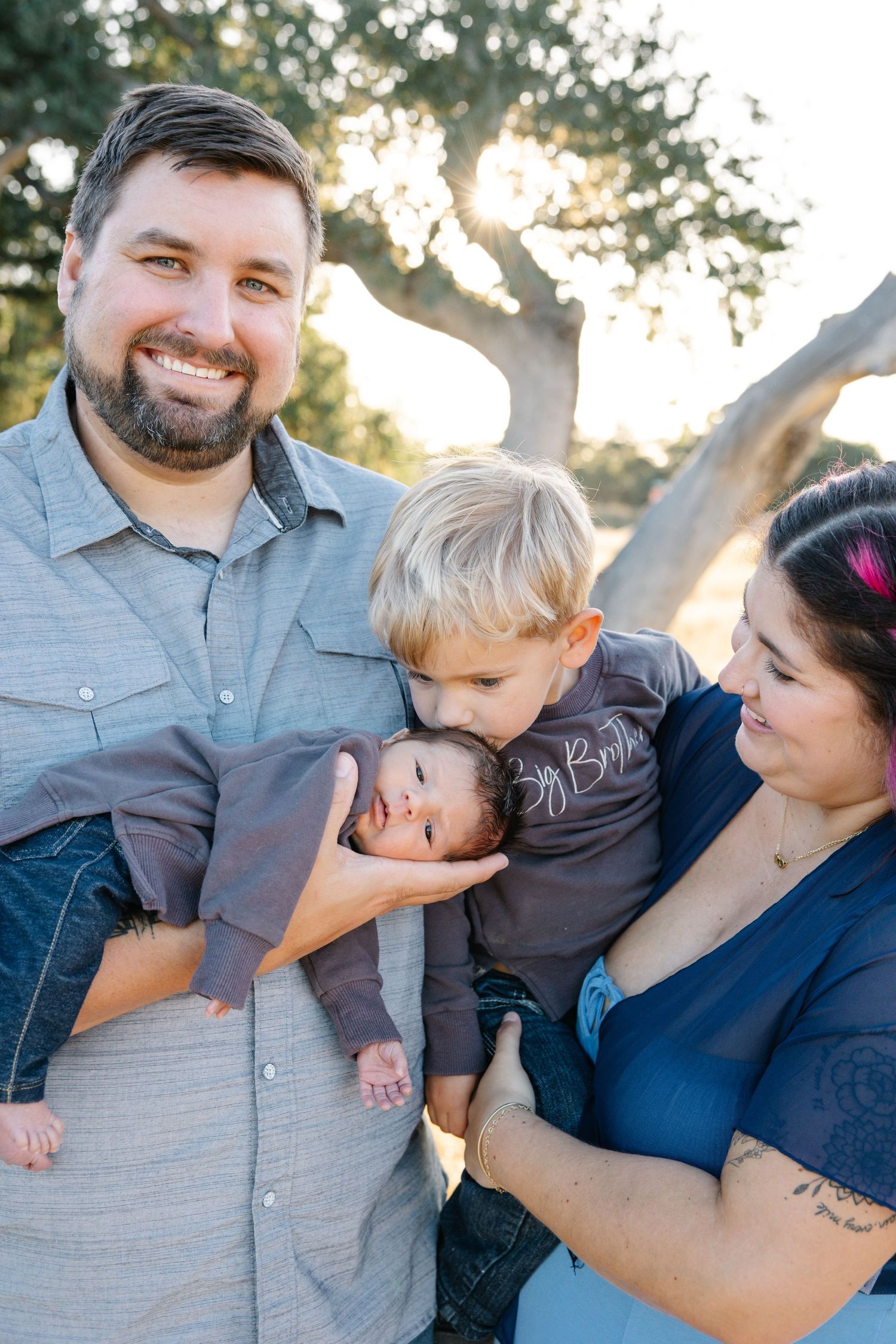 Family of four pose toddler kisses new baby brother on the head while mom embraces in grass and oak trees during sunset, golden hour photo