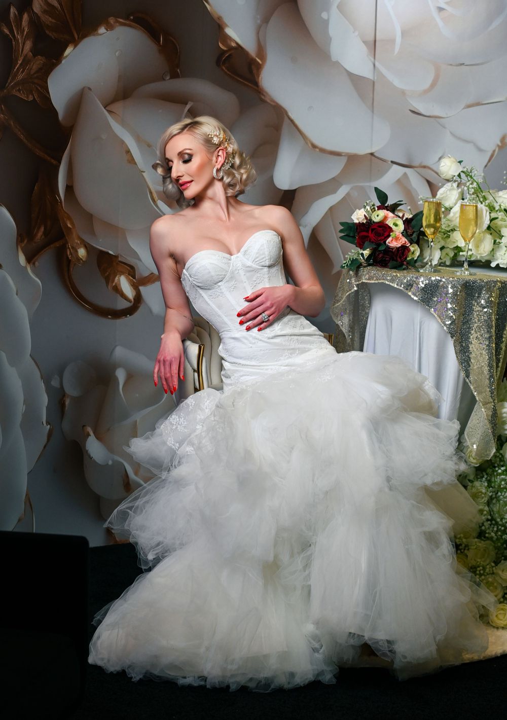 Blonde bride in strapless mermaid wedding gown with ruffled tulle skirt poses elegantly before floral backdrop.