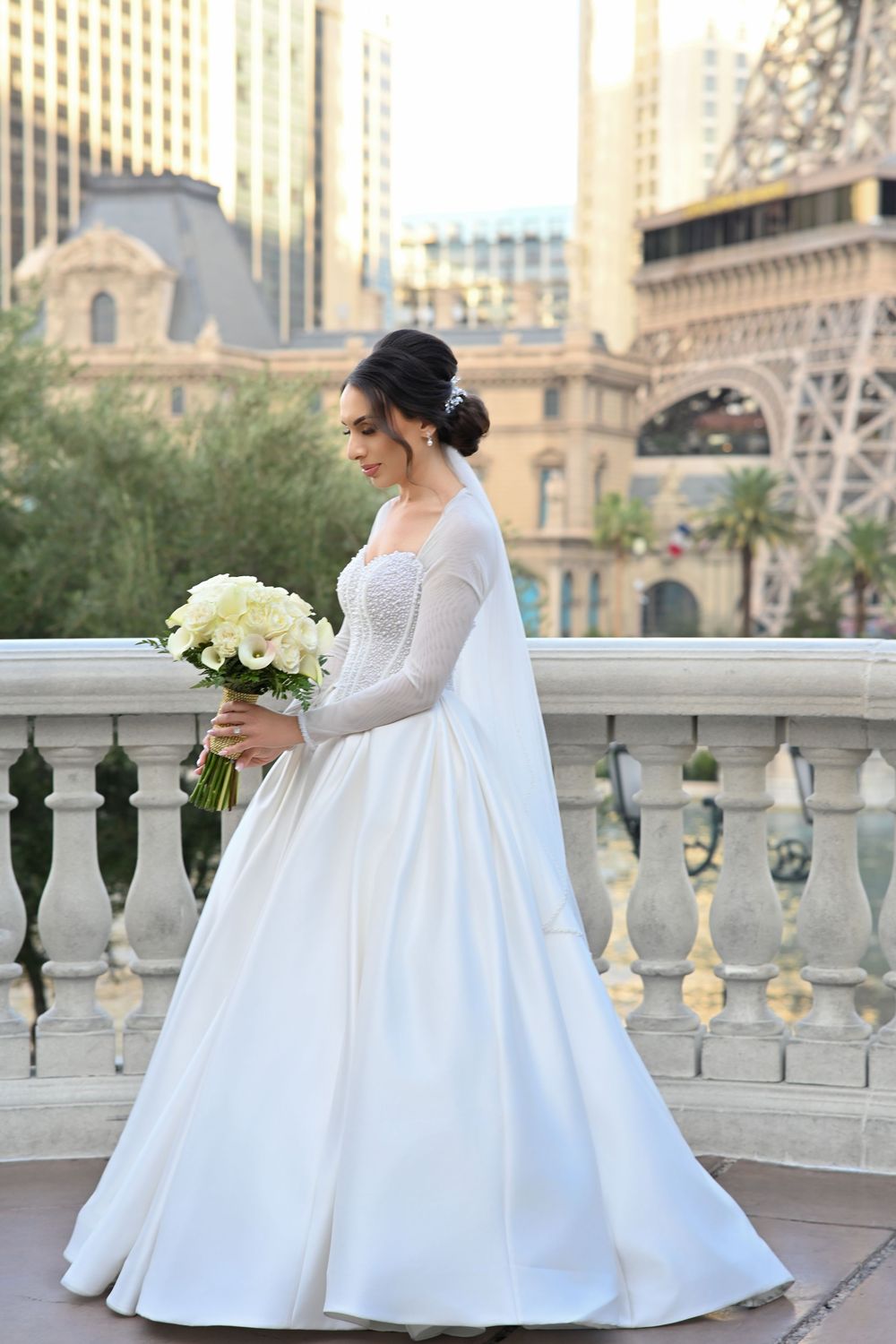 Bride in white long-sleeve ball gown holding white bouquet on balcony with Las Vegas Strip buildings in background.