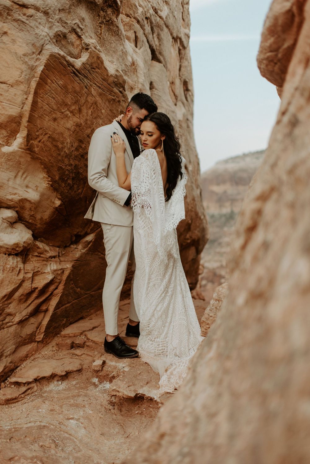Bride and groom embrace among red rock canyon formations, she in lace gown, he in cream suit, desert elopement.