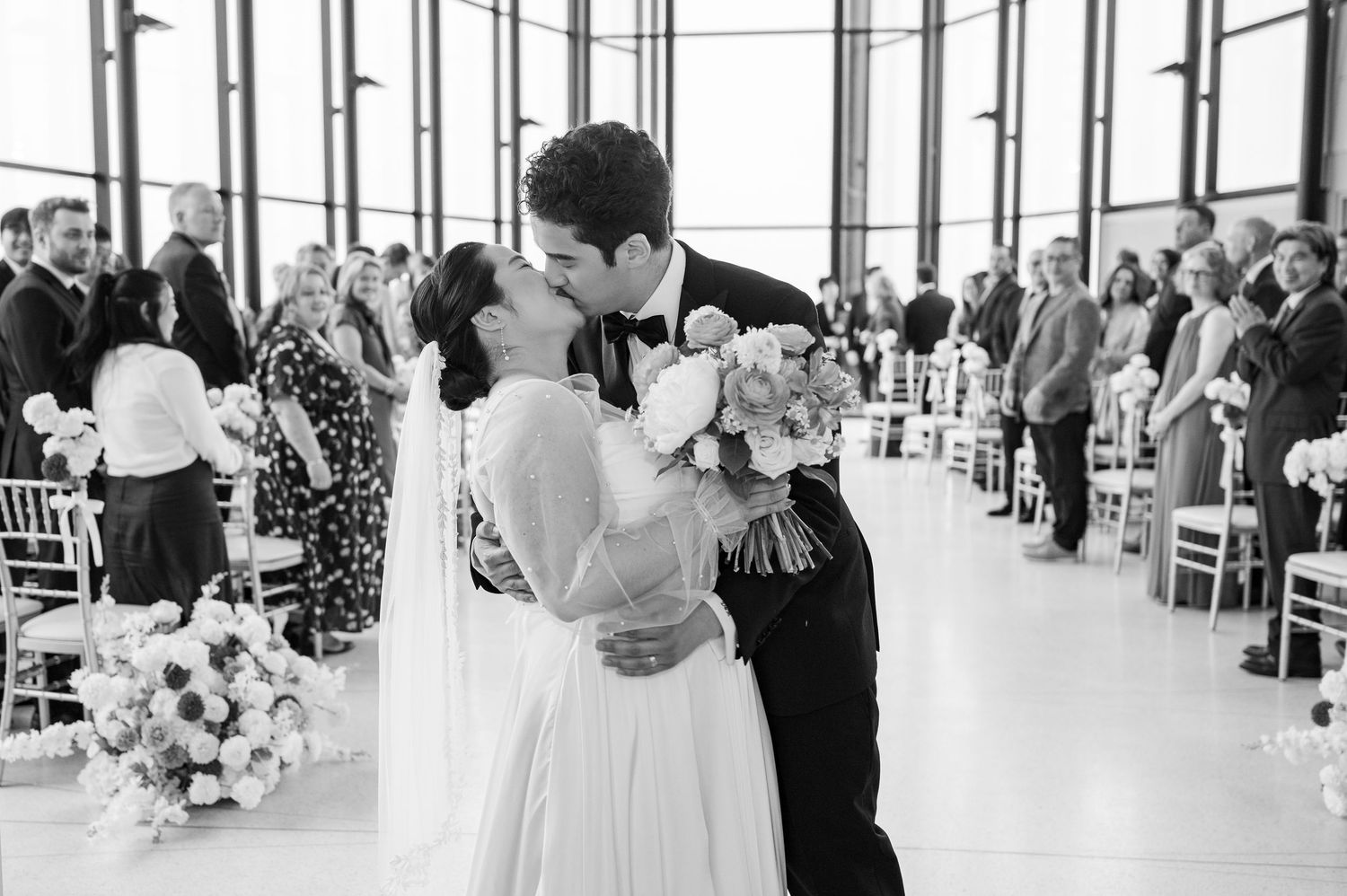 Black and white photo of bride and groom sharing first kiss at elegant wedding ceremony with floral arrangements.